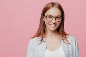 Happy female entrepreneur has straight hair, wears spectacles, smiles positively, enjoys good sales and profits, dressed in stylish shirt, models over pink background with copy space for your text photo