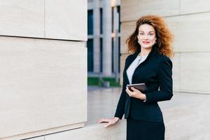 Businesswoman wearing formal clothes, holding modern gadget which she uses for reading messages, e-mails or information. Young female communicating with her partners via digital tablet computer photo