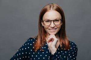 Happy delighted young woman keeps one hand under chin, smiles happily, wears transparent glasses, elegant clohtes, has eyes full of happiness, isolated over white background. Emotions concept photo