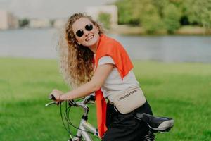 una mujer rubia de cabello rizado va en bicicleta al aire libre, se para con una bicicleta, lleva una bolsa, usa gafas de sol, posa contra el césped verde, usa una camiseta blanca, pantalones negros, suéter rojo en los hombros. estilo de vida al aire libre