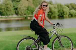 Pleasant looking satisfied curly haired woman spends free time in open air, rides bicycle, spends leisure alone, enjoys freshness of nature, looks at camera with pleasant smile. Summer time. photo