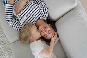 Adopted girl child kiss smiling mother, hugging on cozy sofa at home. Happy motherhood, adoption photo