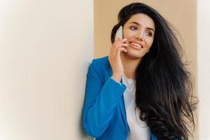 Optimistic female entrepreneur with dark wavy hair, wears makeup, smiles happily, holds modern cell phone near ear has conversation dressed formally looks aside, discusses good news in business sphere photo