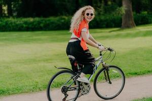 Outdoor image of happy woman rides bike on path, green park around, wears sunglasses, t shirt, pants and sportshoes, has summer holiday, spends spare time actively. People, healthy lifestyle photo