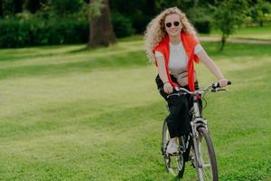 People, nature, rest, lifestyle concept. Happy curly woman rides bicycle among green grass, moves actively, wants to be fit, explores new places in countryside, wears sunglasses, casual clothes photo