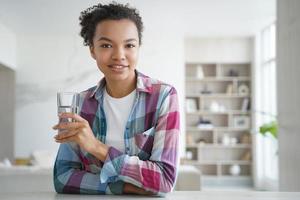 Young mixed race girl holding glass of pure water, sitting at home. Healthy lifestyle, self care photo