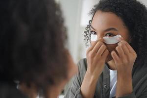 niña afroamericana aplica cosméticos debajo de parches oculares para la piel facial. cuidado de la piel, rutina de belleza