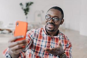 hombre afroamericano con anteojos que sostiene un teléfono inteligente usa aplicaciones modernas que hablan en línea por videollamada foto