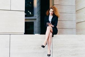 Young female model with curly hair, wearing elegant suit and high-heeled shoes, having slender legs, using modern tablet for communication. Young freelancer working outside, looking seriously photo