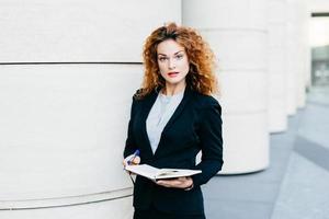 Horizontal portrait of serious pretty businesswoman with curly hair, thin eyebrows and curly hair, wearing black suit and white blouse, holding notebook with pen in hands, looking directly into camera photo