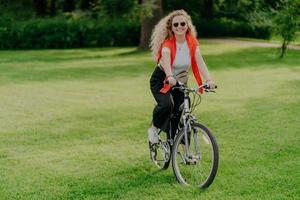 Positive smiling woman dressed in casual clothes, poses on bicycle, has active rest in park, rides on green lawn during sunny summer day, being in high spirit. People, recreation, hobby concept photo