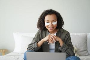 Mixed race girl with under eye patches on face working at laptop in bedroom. Skincare beauty routine photo
