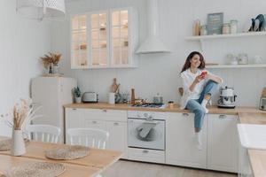 Female holding smartphone shopping online by mobile apps, sitting in modern kitchen interior at home photo