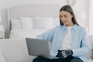 Smiling disabled girl student with bionic hand prosthesis working, learning online at laptop at home photo