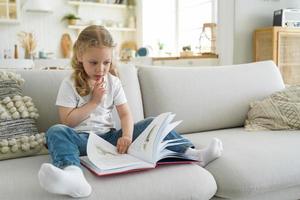 una niña pequeña y pensativa, una niña en edad preescolar, pasa las páginas de un libro de cuentos de hadas para leer, sentarse en un sofá en casa foto