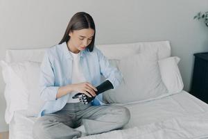 Modern young girl with disability setting her bionic prosthesis of arm sitting on bed at home photo