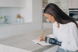 Pretty disabled girl with bionic prosthetic arm writing in notebook sitting at kitchen table at home photo