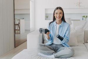 Happy girl with disability enjoying using bionic prosthesis of arm, sitting on sofa at home photo