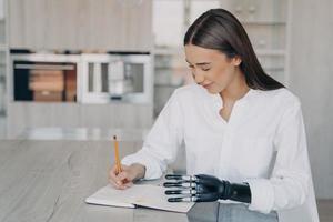 una joven discapacitada sonriente escribiendo notas, sosteniendo un cuaderno con una mano protésica biónica en la mesa foto