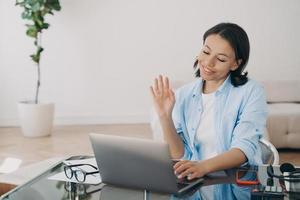 Businesswoman works at laptop, waving hello, having video call with client at office. Remote job photo