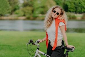 Pretty woman rides bicycle, has bushy blonde hair, stands near her bike, wears sunglasses and white t shirt, enjoys traveling on fresh air, small river or lake in background, green grass. Lifestyle photo