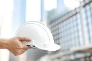 primer plano de la mano de un trabajador de la construcción masculino de ingeniería que sostiene un casco blanco de seguridad para la seguridad de la operación de trabajo. al aire libre del fondo del edificio. foto