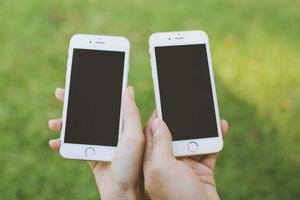 close up of two young male and female  hand holding display white mobile smart phone with blank of empty screen at the outdoors public park. photo