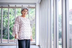 Asian elderly woman using a cane to help her walk near glass window in home, medical care and life insurance concepts. photo