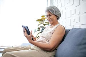 Asian senior woman using smartphone for video call with happy mood smiling and laughing on sofa at home. photo