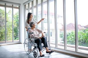 Beautiful asian girl help and take care of senior elderly woman sitting on wheelchair at living room in house, senior therapy patient at home concept. photo