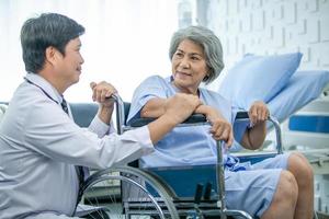 Asian doctor supporting and cheering up senior patient in wheelchair talking, smiling in comfort at hospital. Healthcare and medicine concept. photo