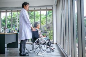 Asian doctor supporting and cheering up senior patient in wheelchair talking, smiling in comfort at home. Healthcare and medicine concept. photo