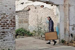 Confident man carrying leather suitcase while walking into ruined building. photo