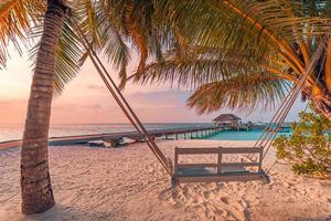 Romantic couple swing close to calm sea and tropical islands shore. Exotic paradise beach landscape, palm trees, sunset sky and summer landscape view. Maldives jetty, water villa