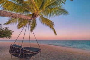 Tropical sunset beach panorama as summer landscape with relax beach swing or hammock on coconut palm, white sand and calm sea beach banner. Perfect beach vacation or summer holiday panoramic concept
