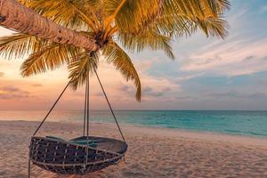 Tropical sunset beach panorama as summer landscape with relax beach swing or hammock on coconut palm, white sand and calm sea beach banner. Perfect beach vacation or summer holiday panoramic concept
