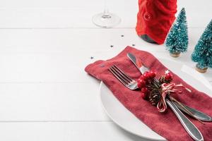 Christmas table setting with white dishware, silverware and red and green decorations on white wooden background photo