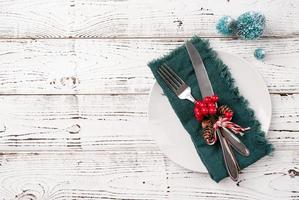 Christmas table setting with white dishware, silverware and red and green decorations on white wooden background. Top view. photo