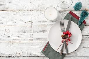 Christmas table setting with white dishware, silverware and red and green decorations on white wooden background. Top view. photo