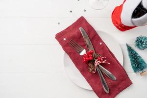 Christmas table setting with white dishware, silverware and red and green decorations on white wooden background. Top view. photo