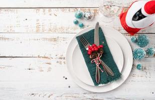 Christmas table setting with white dishware, silverware and red and green decorations on white wooden background. Top view. photo