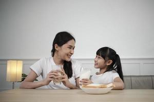 A Healthy Asian Thai family, little daughter, and young mother drink fresh white milk in glass and bread joy together at a dining table in morning, wellness nutrition home breakfast meal lifestyle. photo