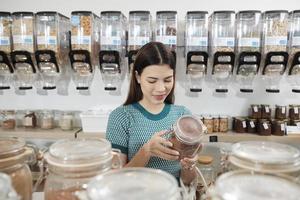 Young Caucasian female customer chooses and shops for organic products in reusable glass jars at refill store, zero-waste grocery, and plastic-free, eco environment-friendly, sustainable lifestyles. photo