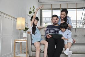 feliz familia tailandesa asiática, padres e hijos se divierten usando una tableta digital juntos en el sofá de la sala de estar, un hermoso fin de semana de ocio y un estilo de vida de bienestar doméstico con tecnología de Internet. foto