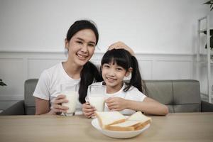 Portrait of healthy Asian Thai family, happy daughter, and young mother looking at camera, drink fresh milk and bread together at dining table in morning, wellness nutrition breakfast meal lifestyle. photo