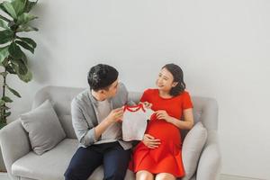 Young husband and a pregnant wife show their baby t-shirt when sitting on sofa. photo