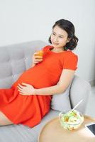 Pretty young Asian pregnant woman eating salad and drinking milk when sitting on sofa. photo