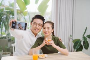 hermosa pareja joven haciendo selfie, usando un teléfono inteligente y sonriendo mientras está sentado en su apartamento foto