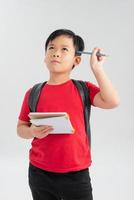 Image of sad smarty boy wearing backpack thinking and touching temple with pen while studying and holding textbooks in hand isolated over white background photo