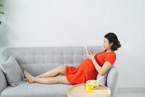 joven embarazada usando el teléfono cuando está acostado en el sofá, comiendo ensalada, bebiendo jugo de naranja. foto